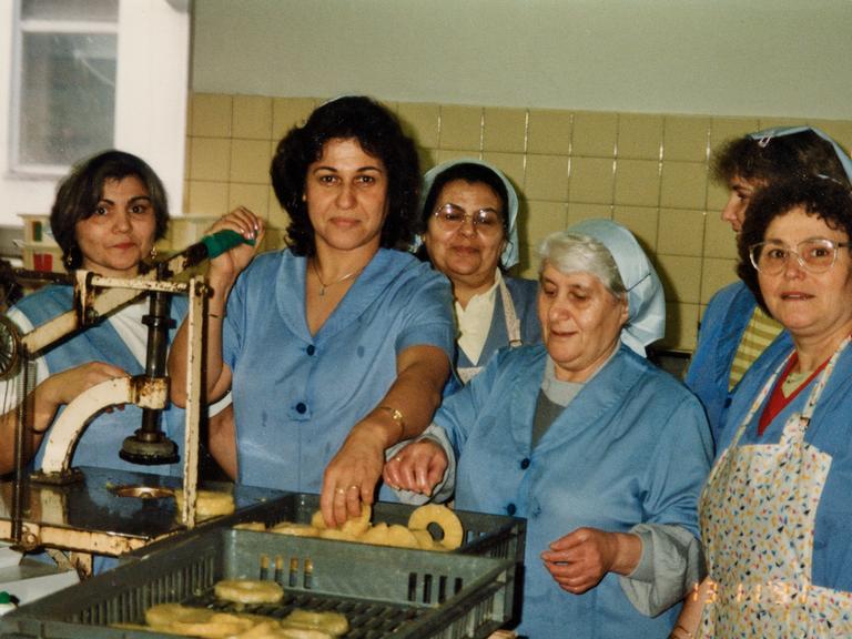 Frauen stehen in blauer Arbeitskleidung an einer Maschine. Arbeiterinnen bei Hachez.