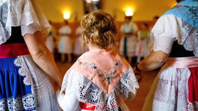 Frauen in sorbischer (wendischer) Tracht singen und tanzen im Dorfgasthof. Im Bild sind zwei Frauen von hinten zu sehen, die zwischen sich ein Kind an den Händen halten. Sie tragen traditionell bestickte Trachten.