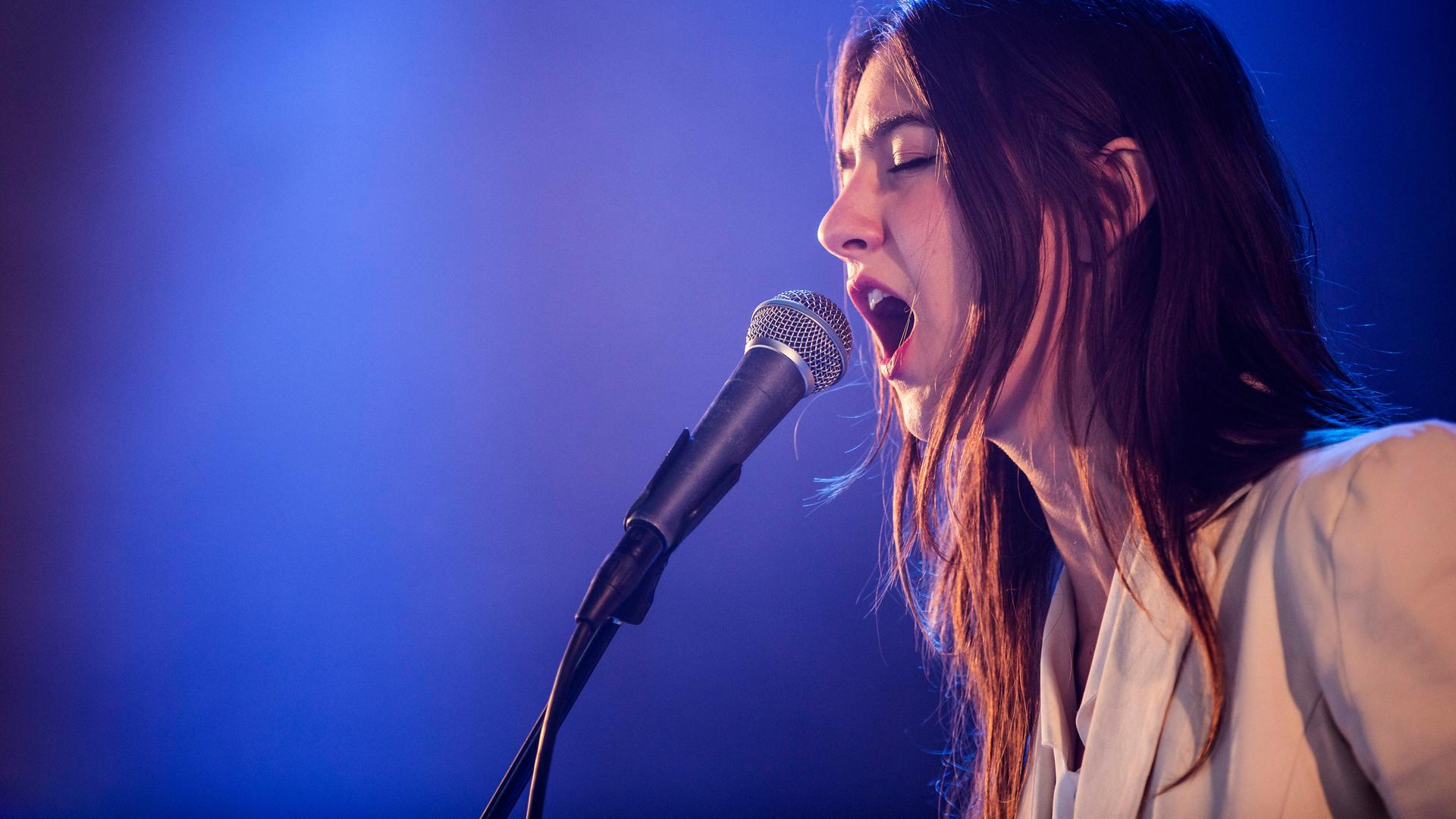 Weyes Blood singt vor blauem Hintergrund leidenschaftlich in ein Mikrofon Weyes Blood singt vor blauem Hintergrund leidenschaftlich in ein Mikrofon