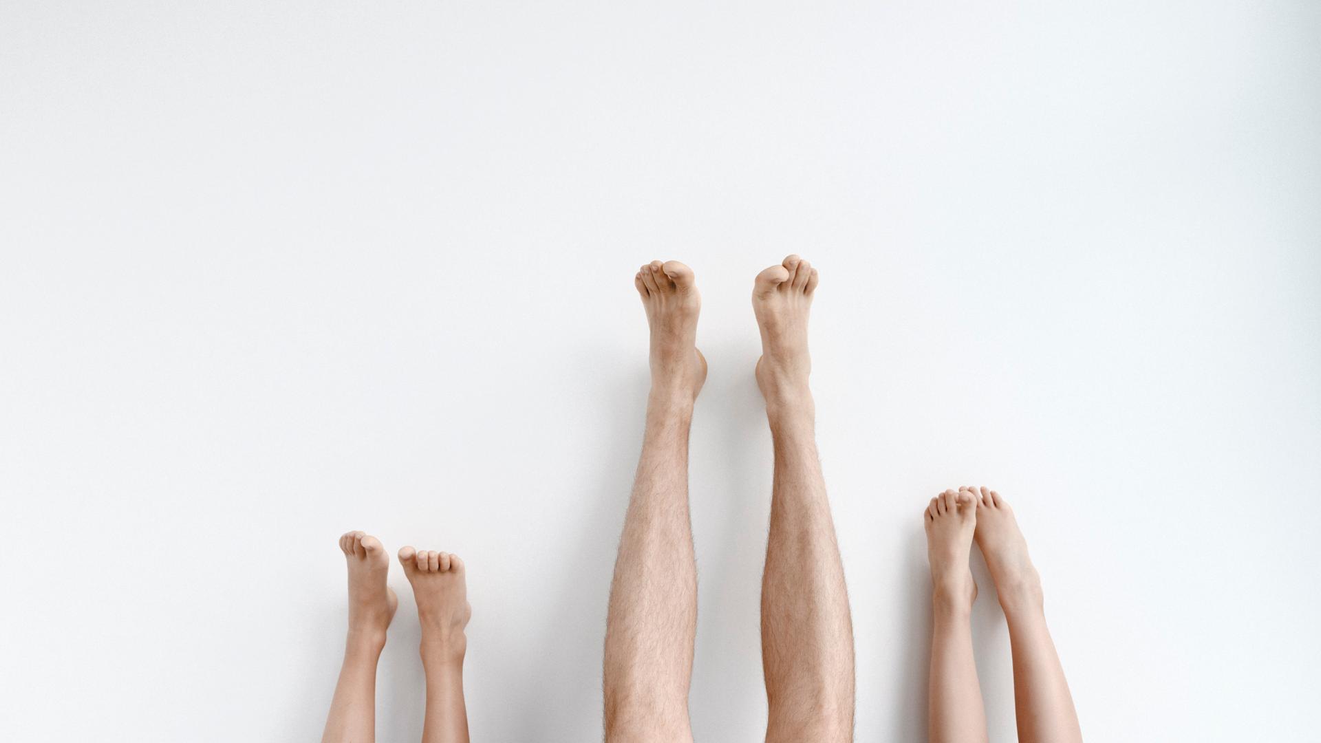 Drei Paar Beine mit den Füßen nach oben an eine Wand gestreckt. Das Paar in der Mitte gehört zu einem Erwachsenen und ist am längsten. Drei Paar Beine mit den Füßen nach oben an eine Wand gestreckt. Das Paar in der Mitte gehört zu einem Erwachsenen und ist am längsten.