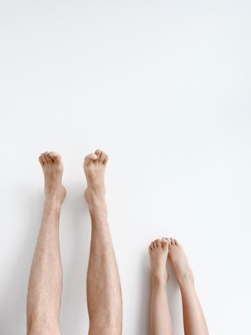 Drei Paar Beine mit den Füßen nach oben an eine Wand gestreckt. Das Paar in der Mitte gehört zu einem Erwachsenen und ist am längsten.