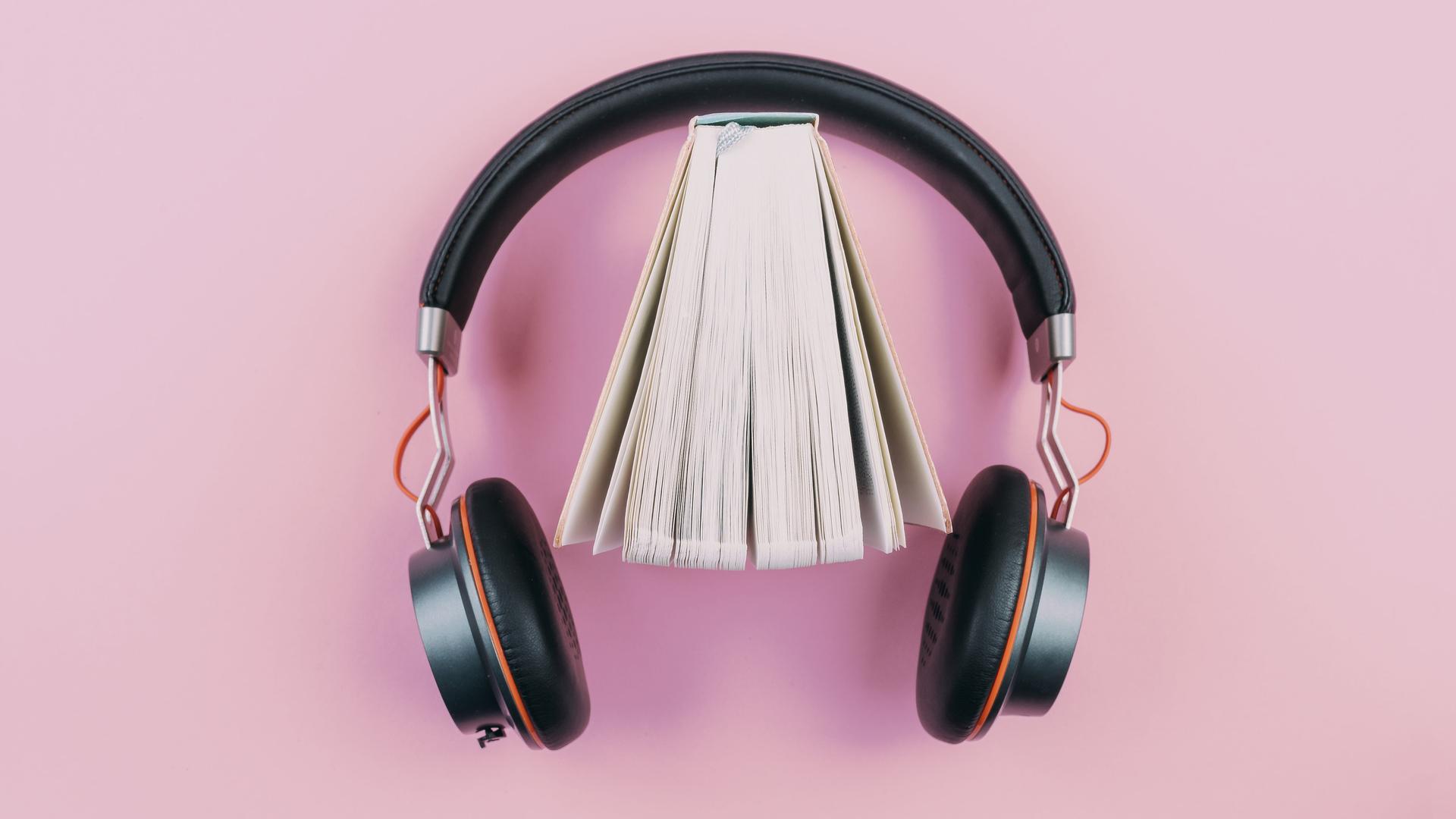 Ein Buch steht leicht aufgefächert in einem Kopfhörer vor rosafarbenem Hintergrund. Ein Buch steht leicht aufgefächert in einem Kopfhörer vor rosafarbenem Hintergrund.