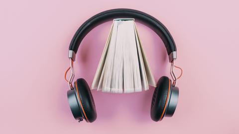 Ein Buch steht leicht aufgefächert in einem Kopfhörer vor rosafarbenem Hintergrund.