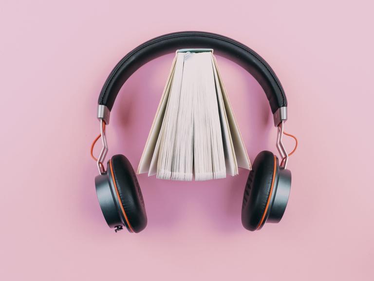 Ein Buch steht leicht aufgefächert in einem Kopfhörer vor rosafarbenem Hintergrund.