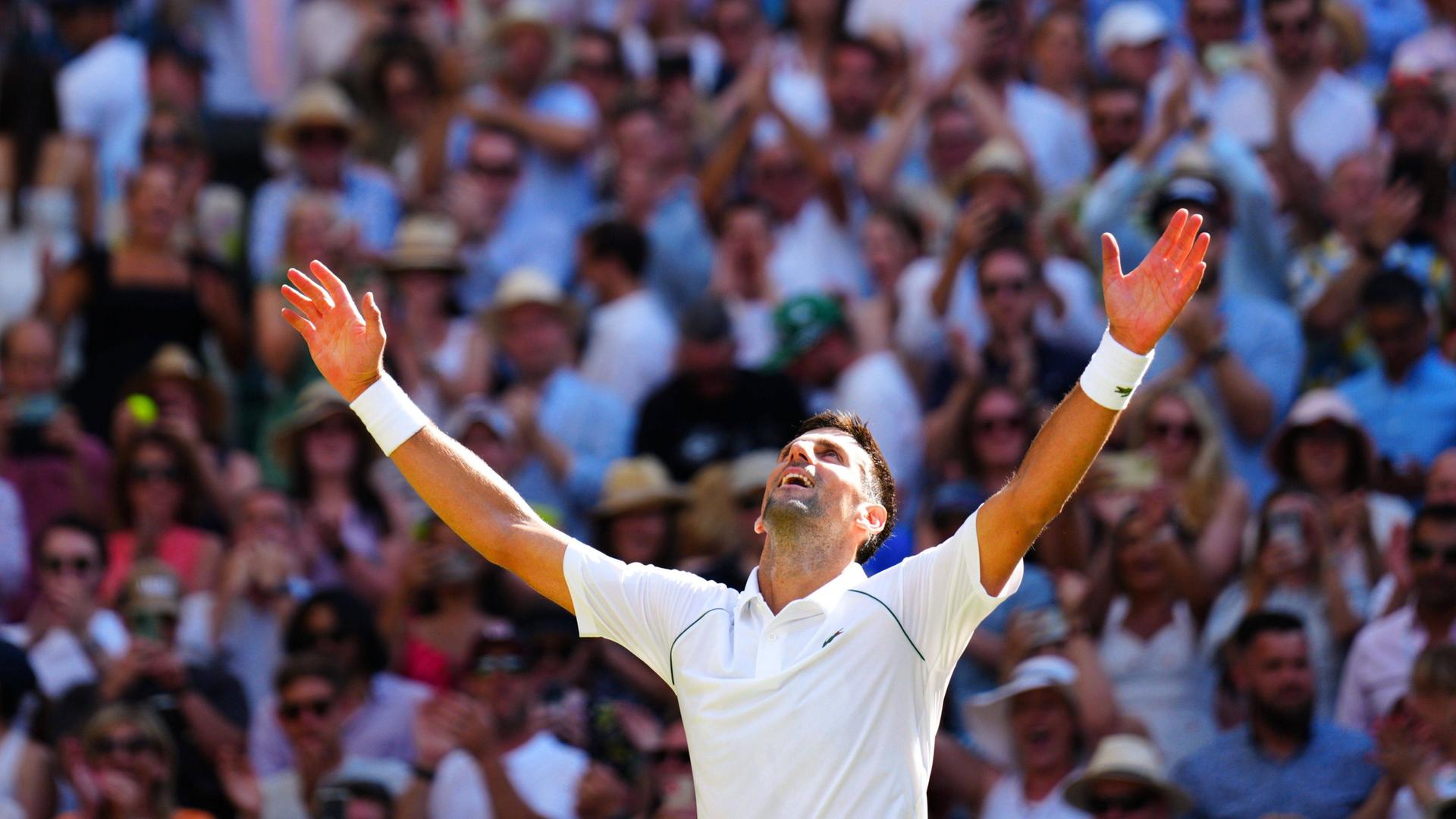 Ein Tennis-Spieler in weißem T-Shirt hebt beide Hände nach oben und jubelt. Im Hintergrund sind zahlreiche Menschen im Publikum zu sehen.