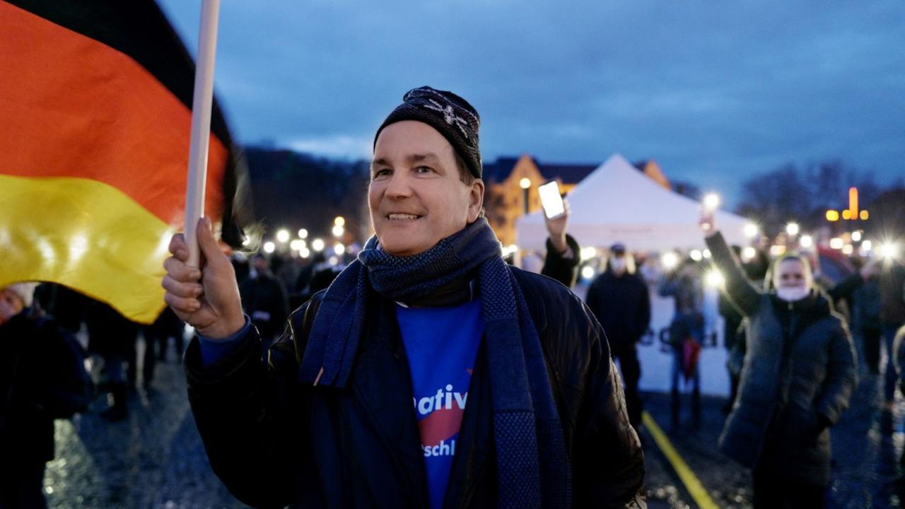 Filmstill aus dem Film "Eine deutsche Partei", Andreas Wild trägt ein blaues AfD-Shirt und schwingt lächelnd eine große, deutsche Flagge bei einer Anti-Corona-Demonstration in Erfurt.