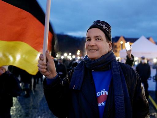 Filmstill aus dem Film "Eine deutsche Partei", Andreas Wild trägt ein blaues AfD-Shirt und schwingt lächelnd eine große, deutsche Flagge bei einer Anti-Corona-Demonstration in Erfurt.