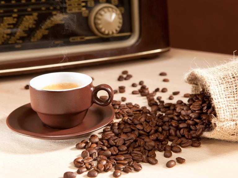 Eine Tasse Kaffee steht vor einem Radiogerät und neben einem umgefallenen Jute-Sack, aus dem Kaffeebohnen gefallen sein.