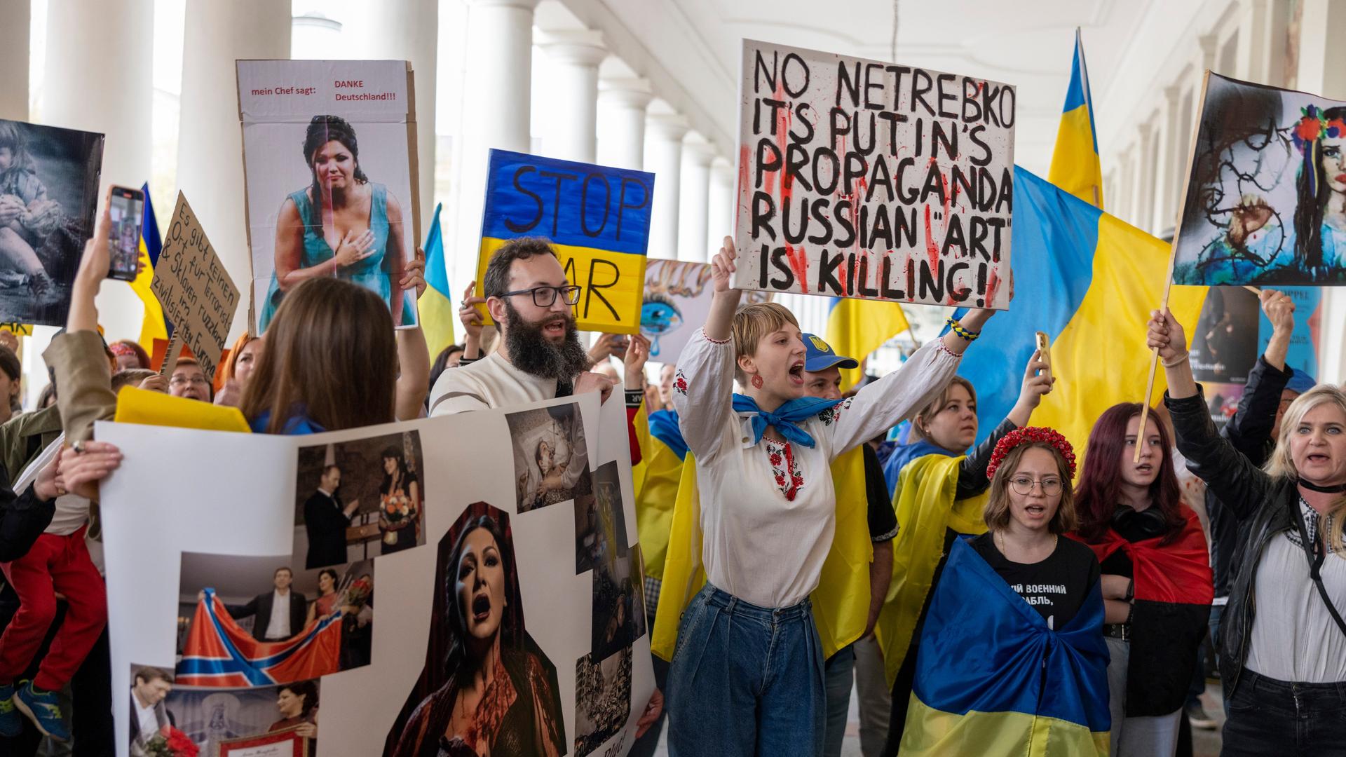 Wir sehen eine Gruppe von Demonstranten, die gegen einen Auftritt der Sopranistin Anna Netrebko protestiert. Sie sind mit Flaggen der Ukraine ausgestattet und halten Plakate gegen Netrebko in die Höhe. Wir sehen eine Gruppe von Demonstranten, die gegen einen Auftritt der Sopranistin Anna Netrebko protestiert. Sie sind mit Flaggen der Ukraine ausgestattet und halten Plakate gegen Netrebko in die Höhe.