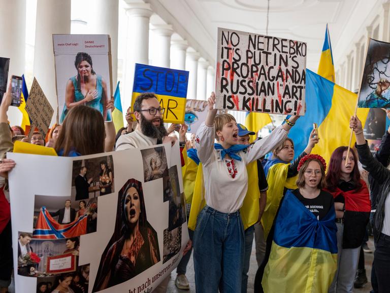 Wir sehen eine Gruppe von Demonstranten, die gegen einen Auftritt der Sopranistin Anna Netrebko protestiert. Sie sind mit Flaggen der Ukraine ausgestattet und halten Plakate gegen Netrebko in die Höhe.