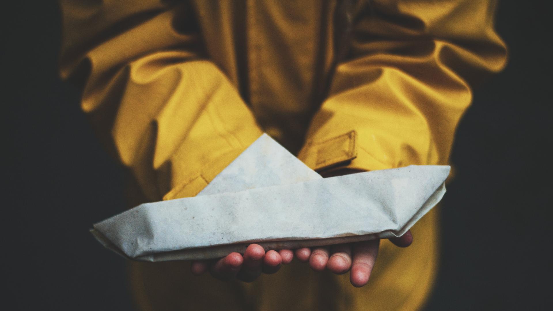 Ein Kind in einem gelben Regenmantel hält ein Papierschiff in Händen. Ein Kind in einem gelben Regenmantel hält ein Papierschiff in Händen.