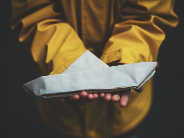 Ein Kind in einem gelben Regenmantel hält ein Papierschiff in Händen.