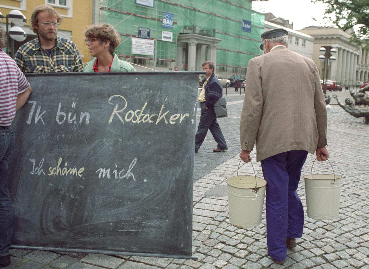Eine Frau hält eine Tafel, auf der geschrieben steht: "Ik bün Rostocker. Ich schäme mich."
