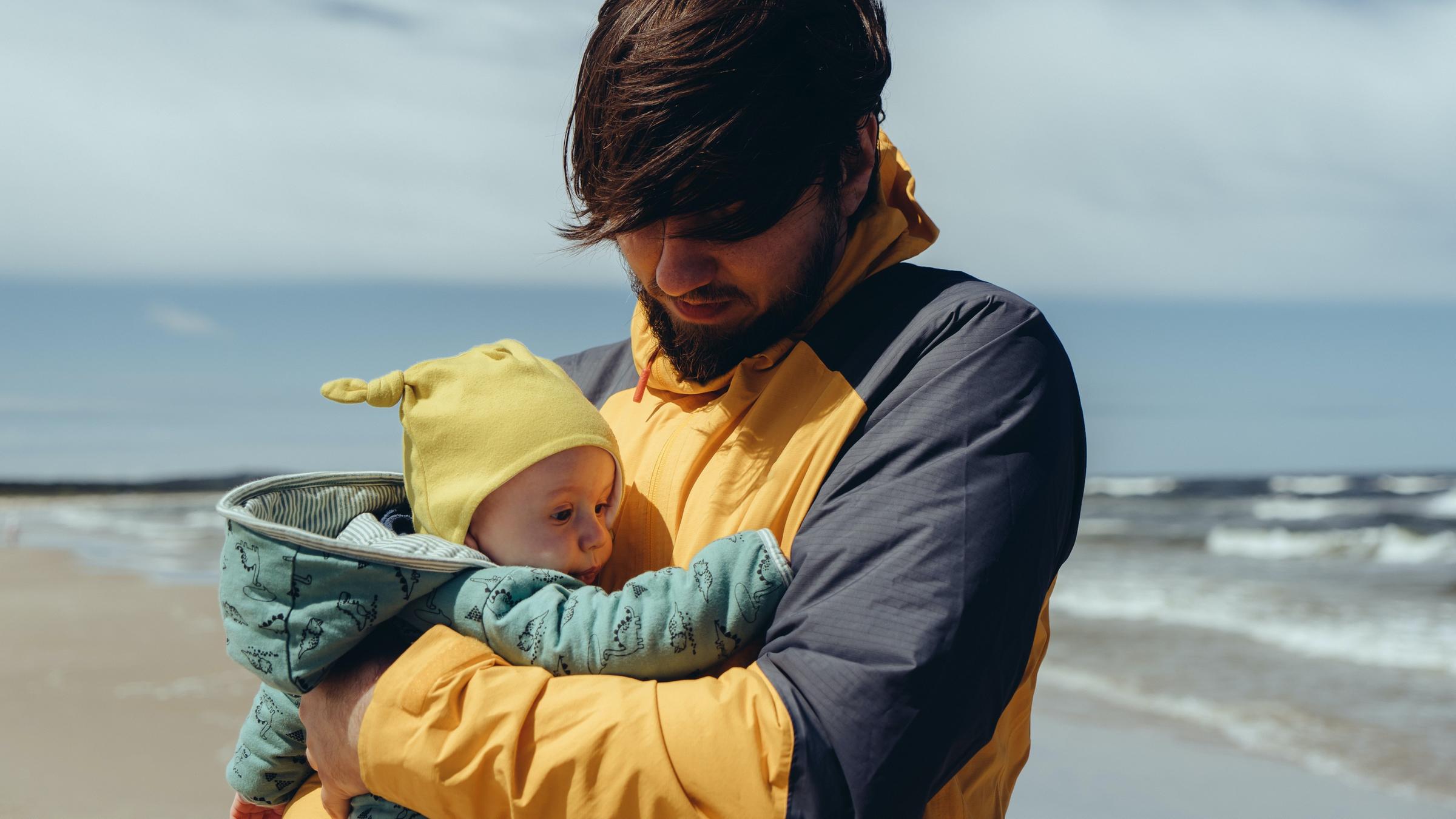 Ein Vater hält am Strand den Säugling eng an seinem Oberkörper.