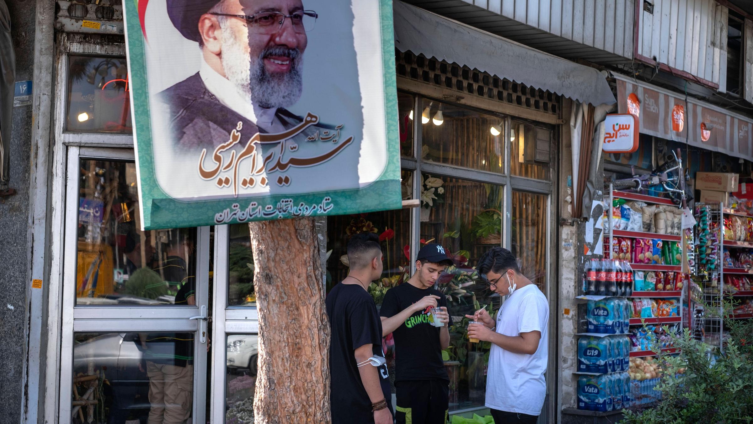Drei Jugendliche stehen vor einem Lebensmittelgeschäft neben einem Baum mit dem Plakat des iranischen Präsidenten Raisi. Drei Jugendliche stehen vor einem Lebensmittelgeschäft neben einem Baum mit dem Plakat des iranischen Präsidenten Raisi.