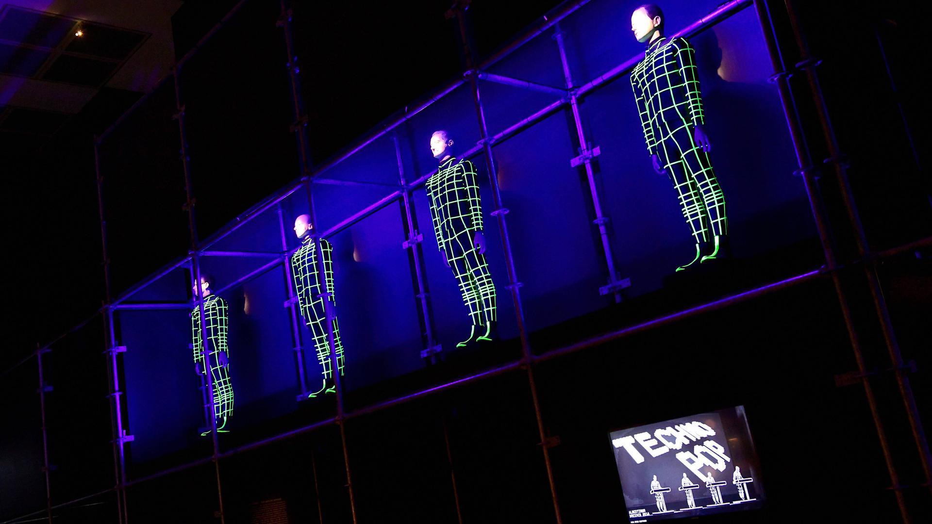 Vier Menschen hängen als elektronische Gitternetze an der Wand. Rechts unten steht auf einem Bildschirm "Techno Pop". Vier Menschen hängen als elektronische Gitternetze an der Wand. Rechts unten steht auf einem Bildschirm "Techno Pop".