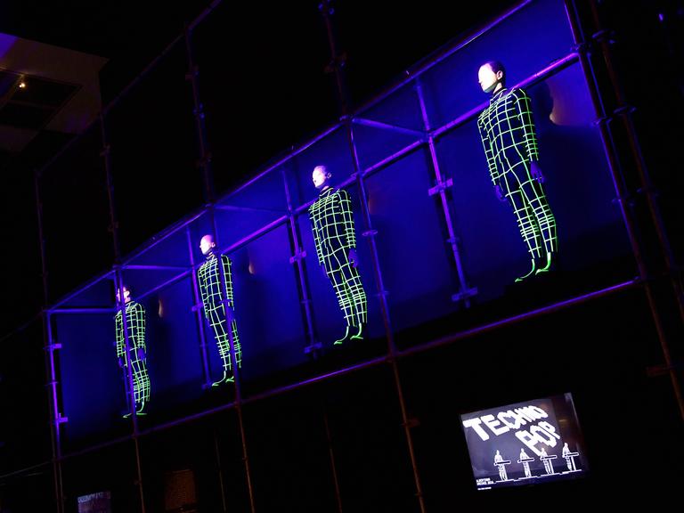Vier Menschen hängen als elektronische Gitternetze an der Wand. Rechts unten steht auf einem Bildschirm "Techno Pop".