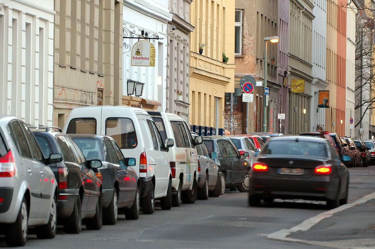 Viele Autos parken am Straßenrand vor Häusern. Auf der Straße fährt ein Auto. Viele Autos parken am Straßenrand vor Häusern. Auf der Straße fährt ein Auto.