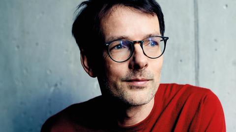 Schriftsteller Jan Böttcher sitzt in einem roten Pullover vor einer Beton-Wand
