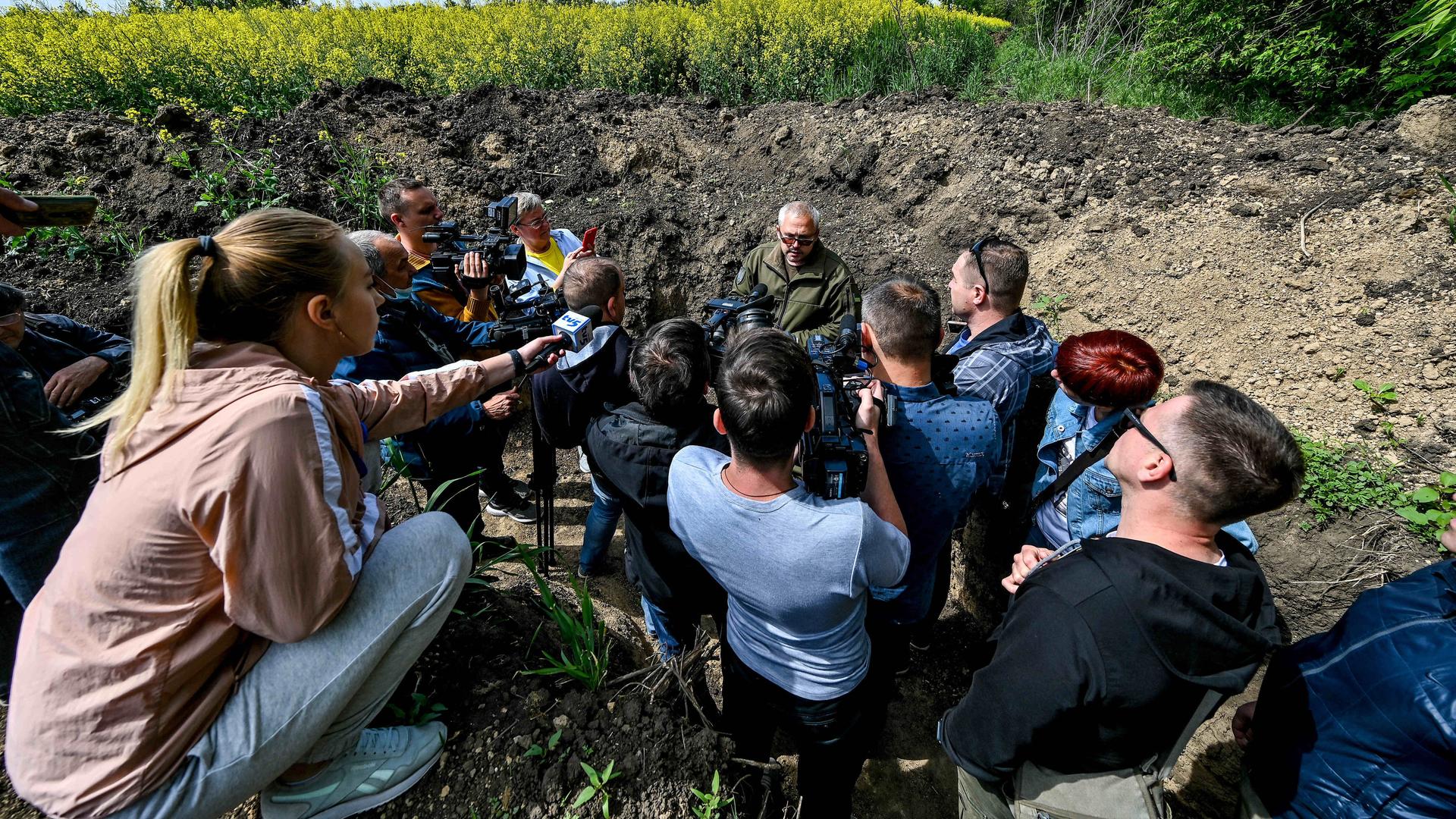 Medienvertreter sitzen im Kreis um einen ukrainischen Militär im Feld. 