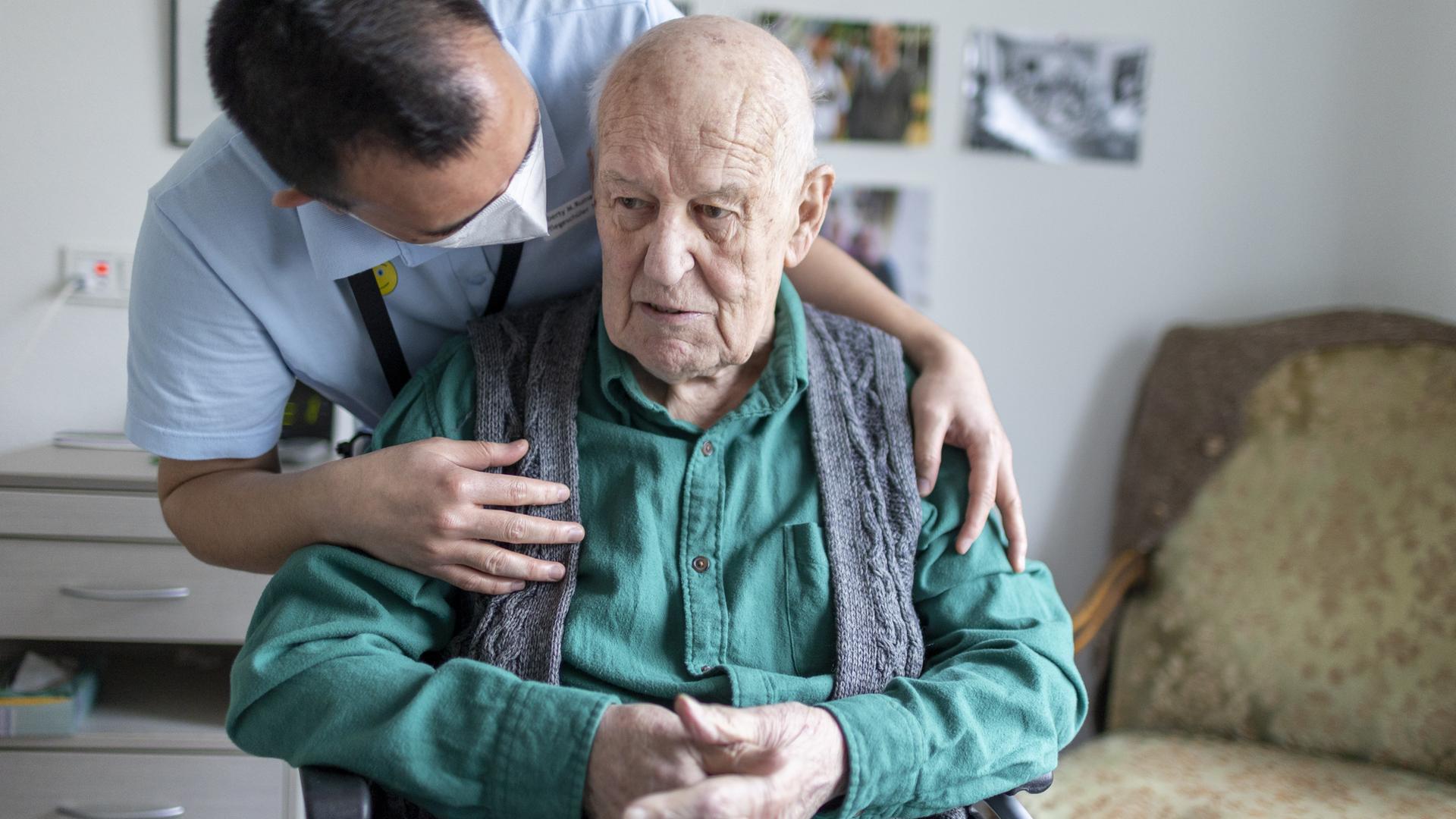 Ein Altenpfleger beugt sich über die Schulter eines alten, sitzenden Mannes und wendet sich ihm zu.