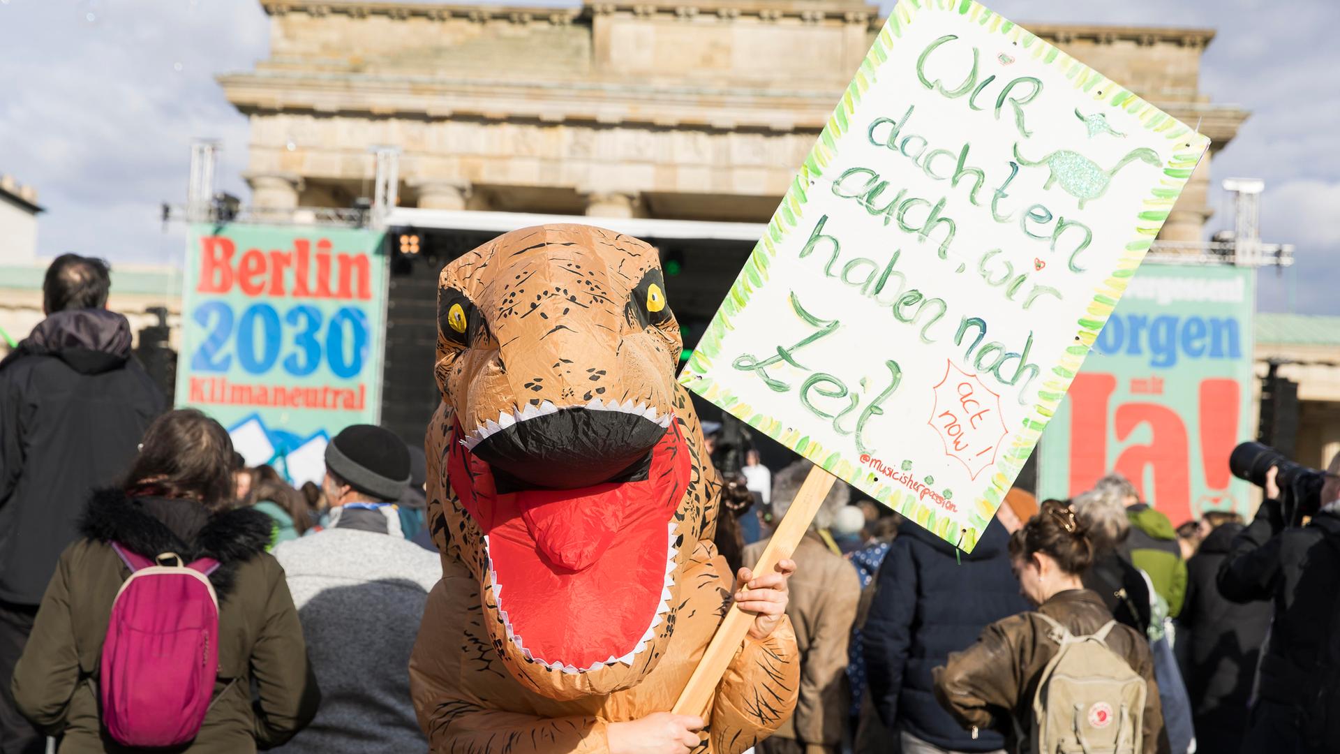 Teilnehmerin im Dinosaurier-Kostüm mit Schild "Wir dachten auch, wir haben noch Zeit" bei der Berlin Climate Aid Demo am Brandenburger Tor.