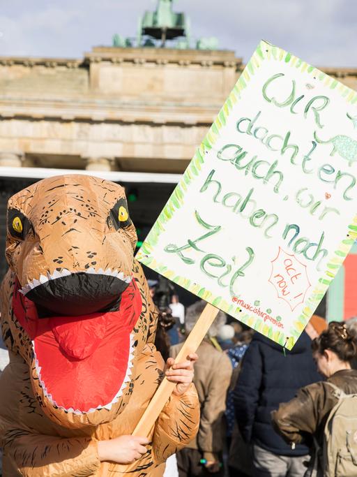 Teilnehmerin im Dinosaurier-Kostüm mit Schild "Wir dachten auch, wir haben noch Zeit" bei der Berlin Climate Aid Demo am Brandenburger Tor.