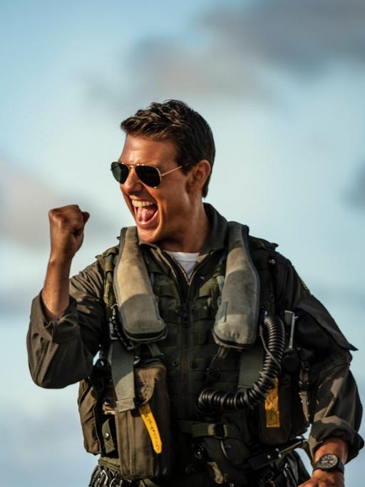 Tom Cruise in der Rolle des Capt. Pete Maverick Mitchell im Film "Top Gun: Maverick" von Joseph Kosinski. Er trägt einen Pilotenanzug und eine dunkle Sonenbrille und ballt die Faust jubelnd.