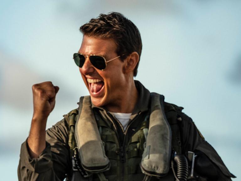 Tom Cruise in der Rolle des Capt. Pete Maverick Mitchell im Film "Top Gun: Maverick" von Joseph Kosinski. Er trägt einen Pilotenanzug und eine dunkle Sonenbrille und ballt die Faust jubelnd.