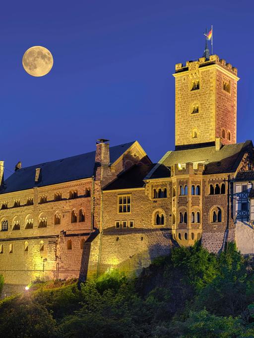 Die alten Mauern der Wartburg sind nachts erleuchtet, während ein Vollmond direkt über der Burg steht.