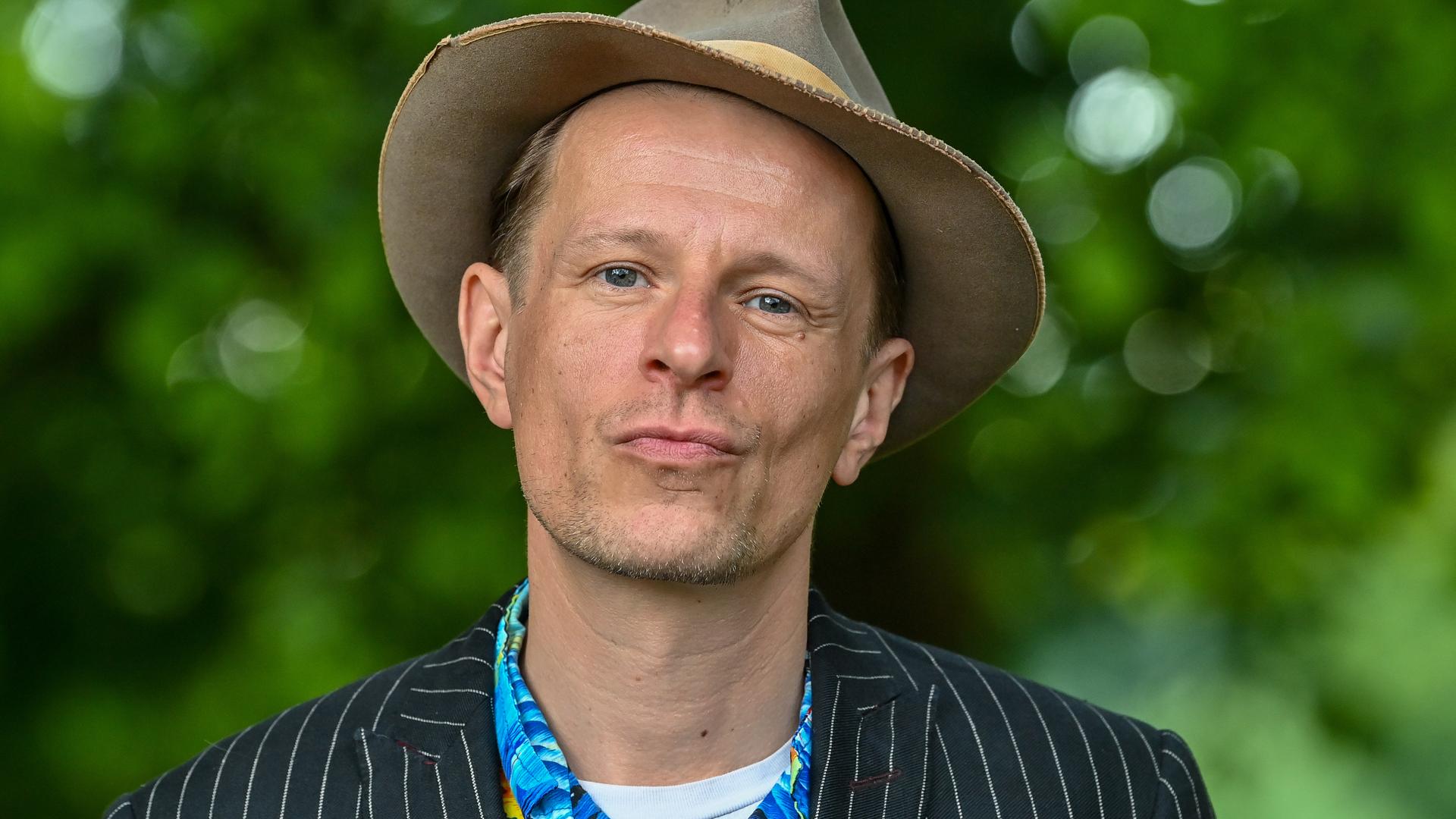 Der Schauspieler Alexander Scheer schaut in die Kamera, im Hintergrund leuchtet das Sommergrün eines Parks. Der Schauspieler Alexander Scheer schaut in die Kamera, im Hintergrund leuchtet das Sommergrün eines Parks.