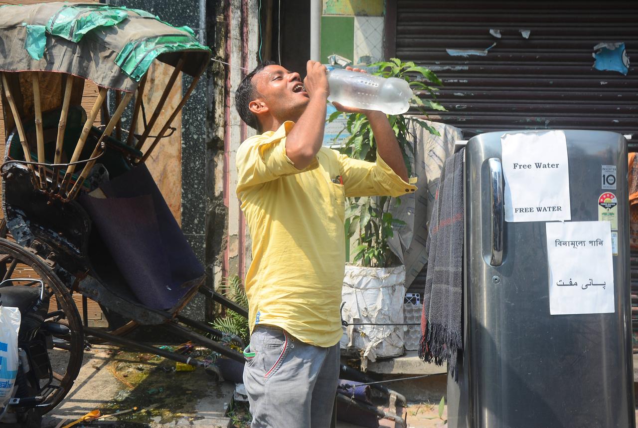 Ein Mann mit gelbem T-Shirt setzt eine volle Wasserflasche an den Mund und lehnt den Kopf zurück. Im Hintergrund eine Rikscha.