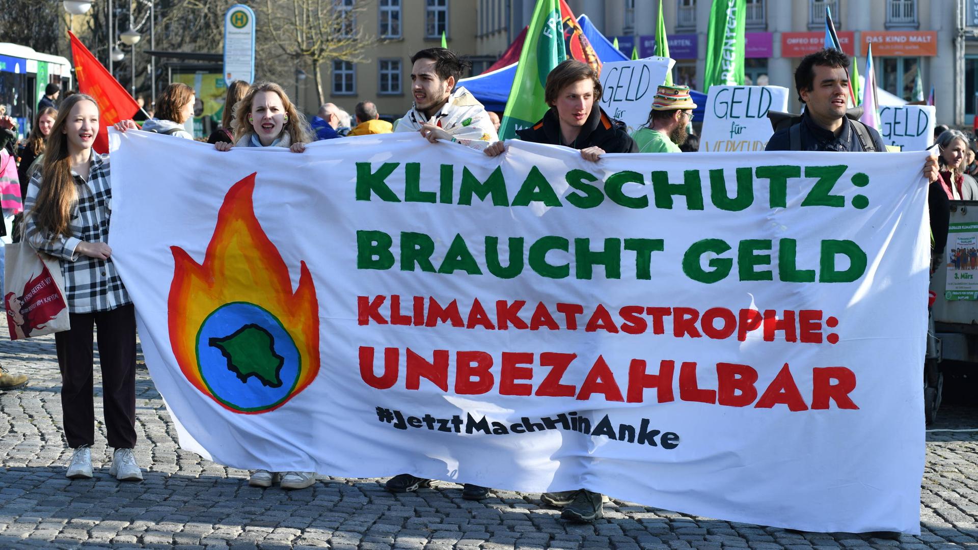 Zwei junge Frauen und drei junge Männer halten ein Transparent auf dem steht: "Klimaschutz braucht Geld. Klimakatastrophe: unbezahlbar. Jetzt mach hin, Anke." Zwei junge Frauen und drei junge Männer halten ein Transparent auf dem steht: "Klimaschutz braucht Geld. Klimakatastrophe: unbezahlbar. Jetzt mach hin, Anke."
