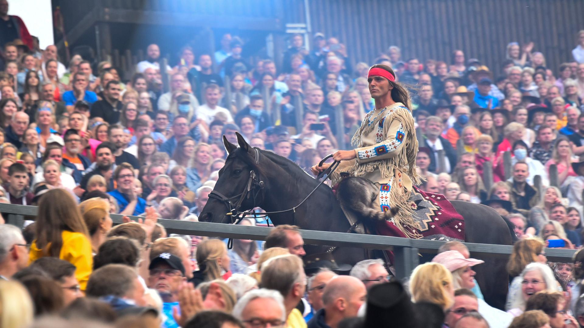 Ein Mann auf einen Pferd mit Federschmuck reitet durch eine Menschenmenge: Aufführung von "Der Ölprinz" bei den Karl May Spielen 2022 in Bad Segeberg. Ein Mann auf einen Pferd mit Federschmuck reitet durch eine Menschenmenge: Aufführung von "Der Ölprinz" bei den Karl May Spielen 2022 in Bad Segeberg.