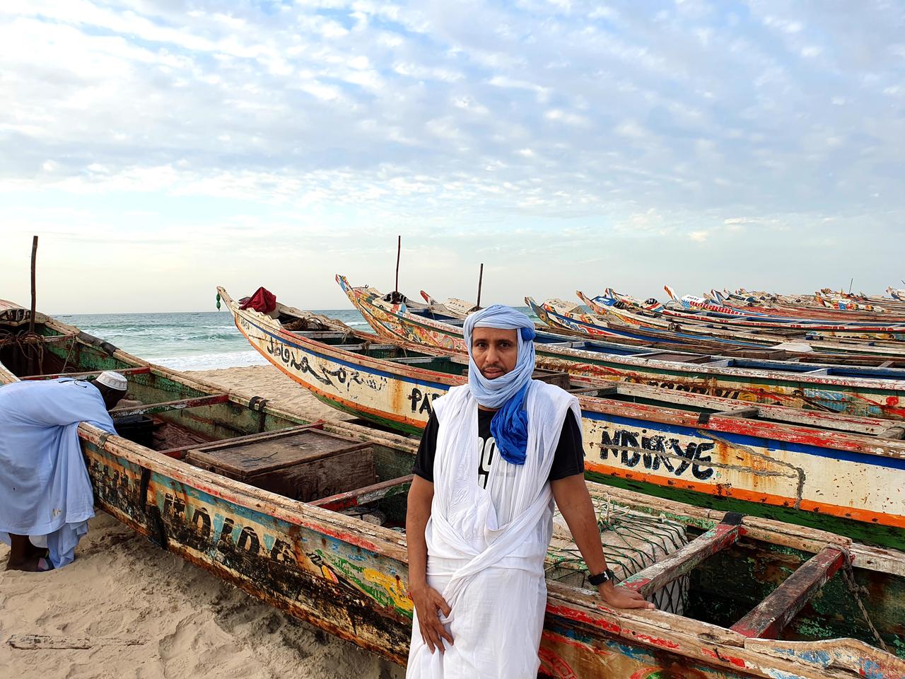 Mohamedou Slahi steht mit Kopftuch bekleidet zwischen bunten Booten am Strand