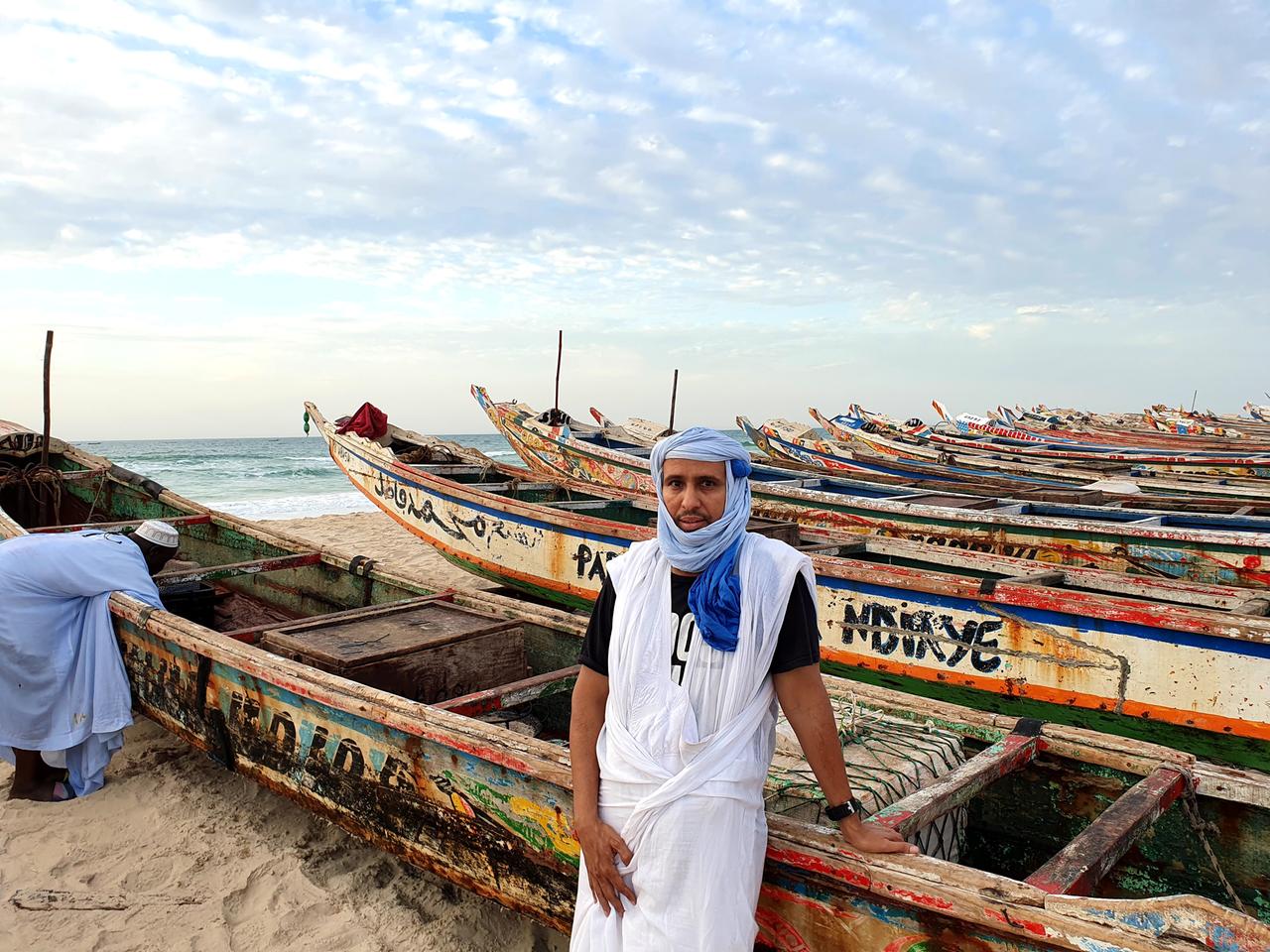Mohamedou Slahi steht mit Kopftuch bekleidet zwischen bunten Booten am Strand Mohamedou Slahi steht mit Kopftuch bekleidet zwischen bunten Booten am Strand