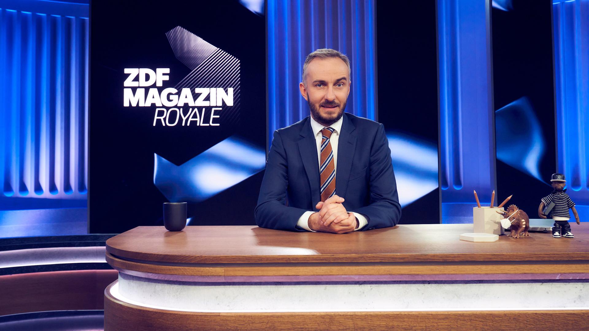 Jan Böhmermann sitzt an einem Tisch im Studio und schaut in die Kamera.