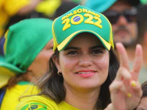 Eine hübsche junge Frau in gelbem Shirt trägt ein grünes Käppi auf dem "Bolsonaro 2022" steht.