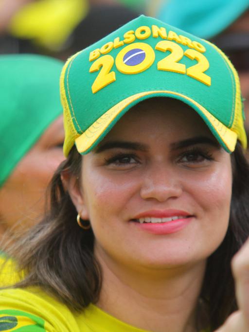 Eine hübsche junge Frau in gelbem Shirt trägt ein grünes Käppi auf dem "Bolsonaro 2022" steht.