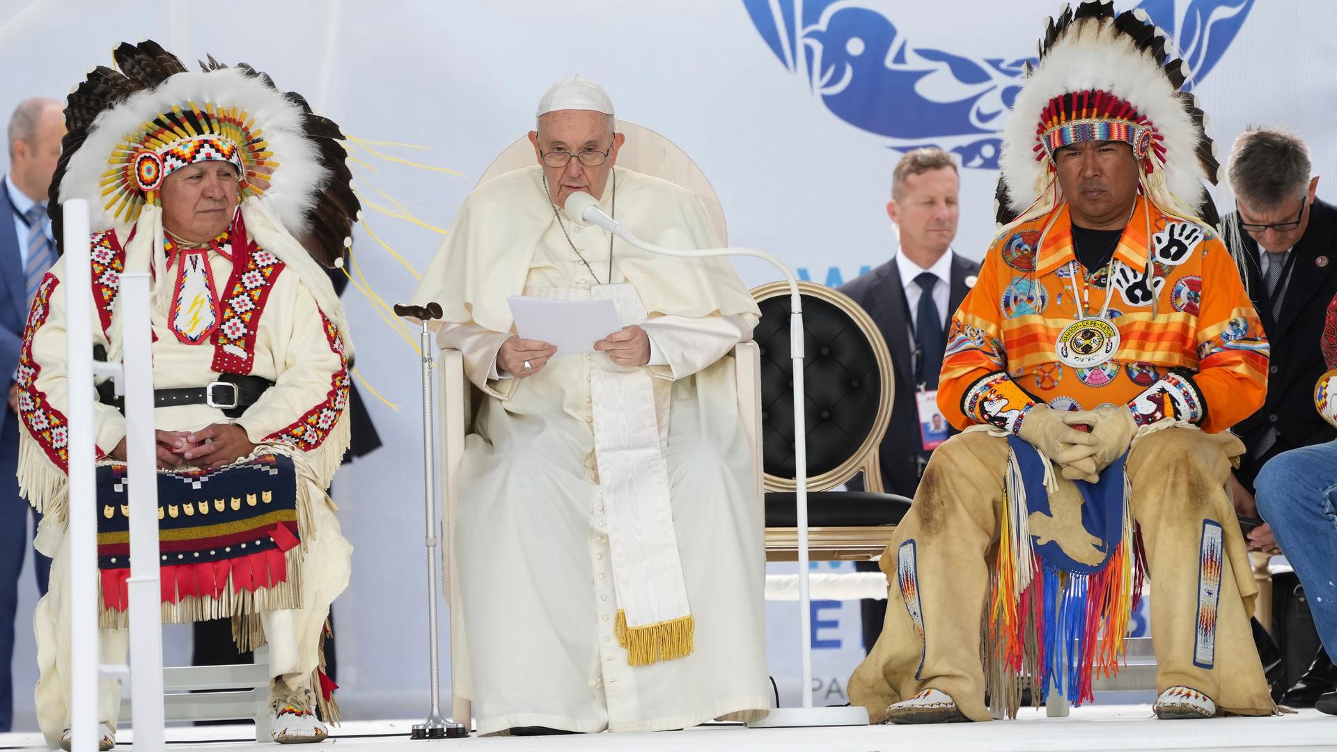 Papst Franziskus spricht umrahmt von zwei Indigenen-Vertretern in ein Mikrofon.