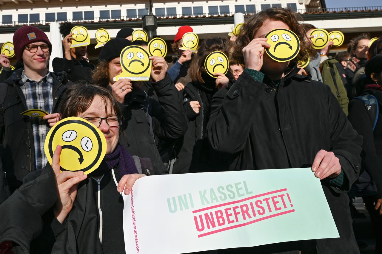 Menschen mit traurigen Smileys vor den Gesichtern udn einem Schild: "Uni Kassel unbefristet". Menschen mit traurigen Smileys vor den Gesichtern udn einem Schild: "Uni Kassel unbefristet".