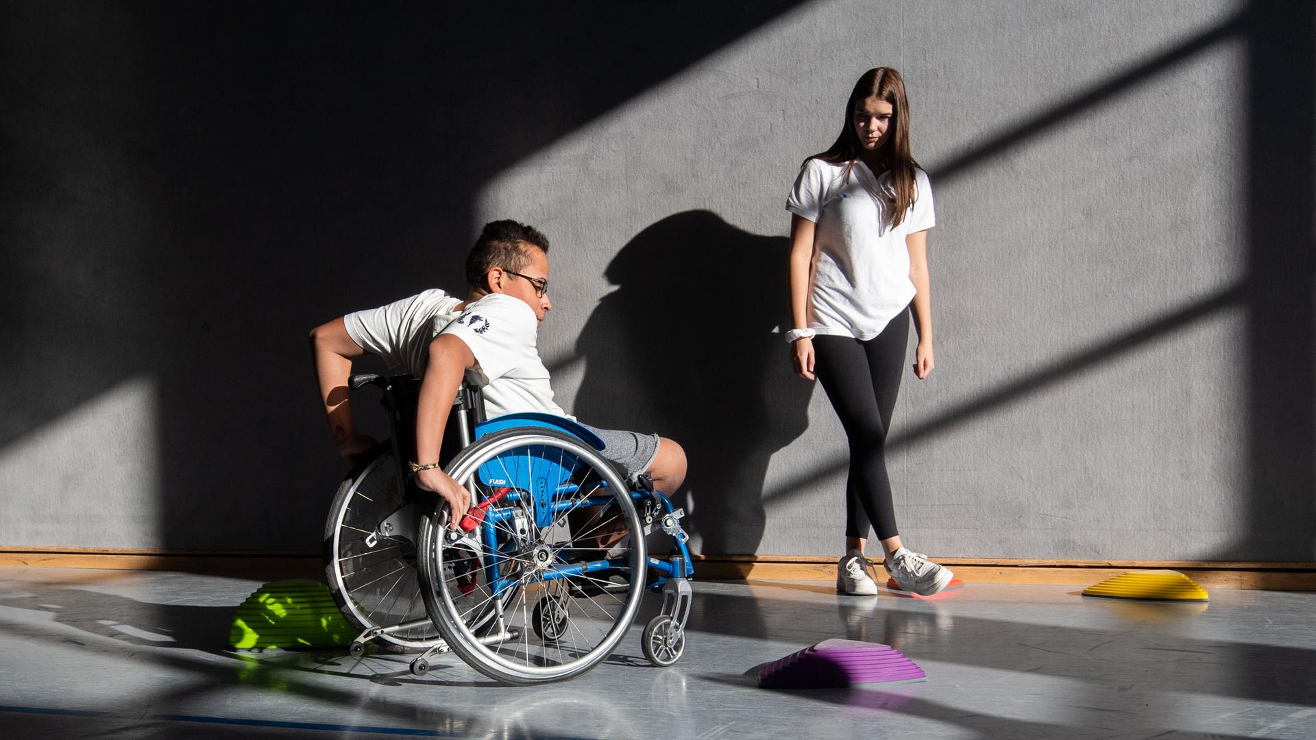 Ein Schüler der Förderschule Paul-Klee-Schule in Celle fährt mit seinem Rollstuhl in der Sporthalle des Gymnasium Burgdorf Slalom um Hindernisse, während eine Schülerin zuschaut.
