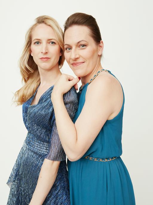 Zwei junge Frauen stehen nebeneinander, blicken freundlich in die Kamera. Die linke Frau hat blonde lange Haare, die rechte Frau hat dunkelbraune Haare, zurückgesteckt.