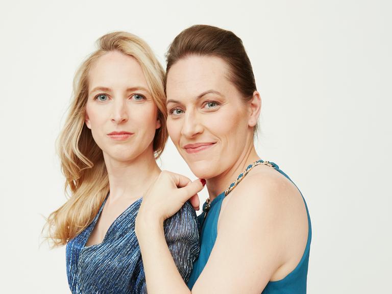 Zwei junge Frauen stehen nebeneinander, blicken freundlich in die Kamera. Die linke Frau hat blonde lange Haare, die rechte Frau hat dunkelbraune Haare, zurückgesteckt.