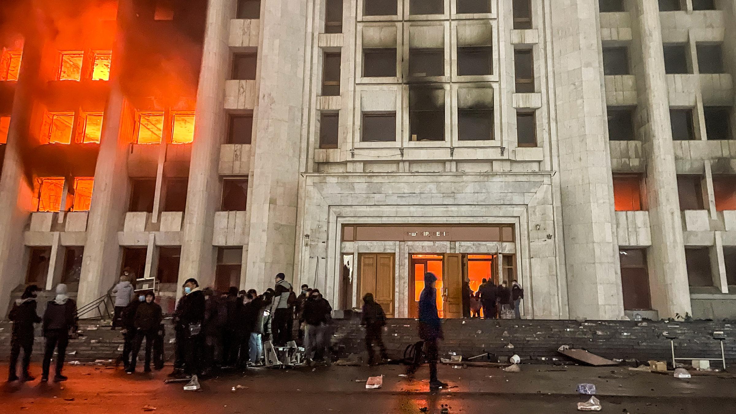 Schwarz gekleidete Menschen stehen vor dem Büro des Bürgermeisters in Almaty, Kasachstan. Das Gebäude steht in Flammen. 