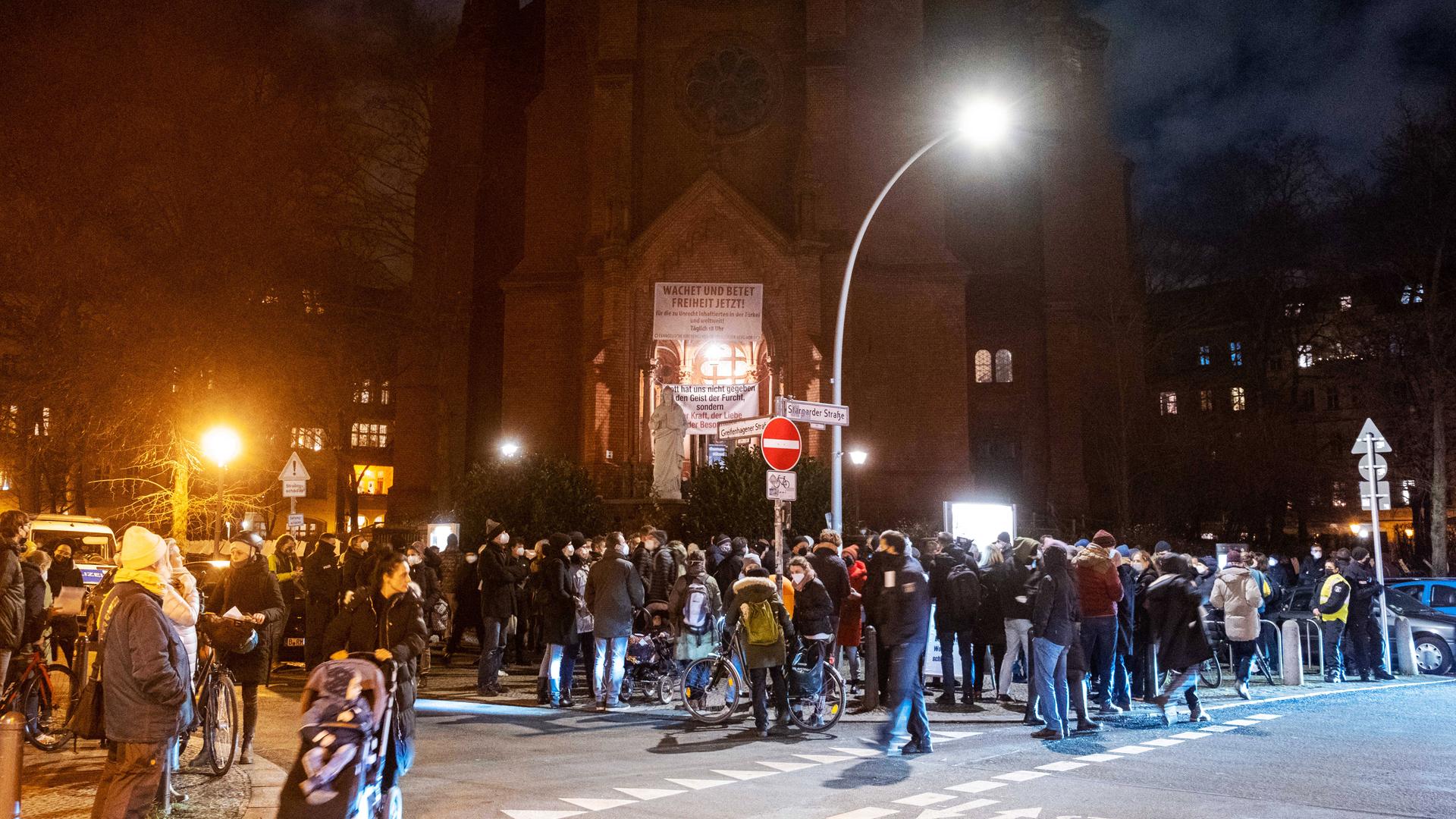 Viele Menschen mit Mundschutz stehen am Abend auf der Straße, vor dem Eingang einer Backsteinkirche.