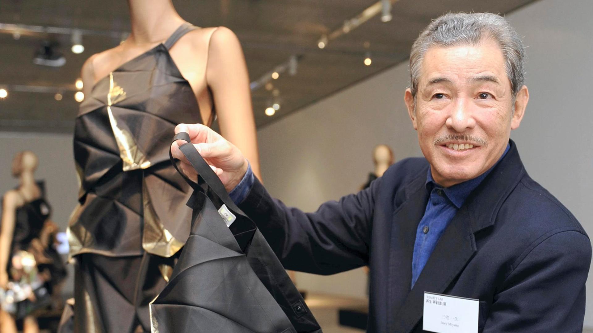 Der japanische Modedesigner Issey Miyake hält ein Stück Stoff in die Kamera. Daneben steht eine Schneiderpuppe, die einen Entwurf von ihm trägt. Miyake trägt einen dunklen Anzug. Der japanische Modedesigner Issey Miyake hält ein Stück Stoff in die Kamera. Daneben steht eine Schneiderpuppe, die einen Entwurf von ihm trägt. Miyake trägt einen dunklen Anzug.
