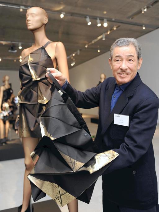 Der japanische Modedesigner Issey Miyake hält ein Stück Stoff in die Kamera. Daneben steht eine Schneiderpuppe, die einen Entwurf von ihm trägt. Miyake trägt einen dunklen Anzug.