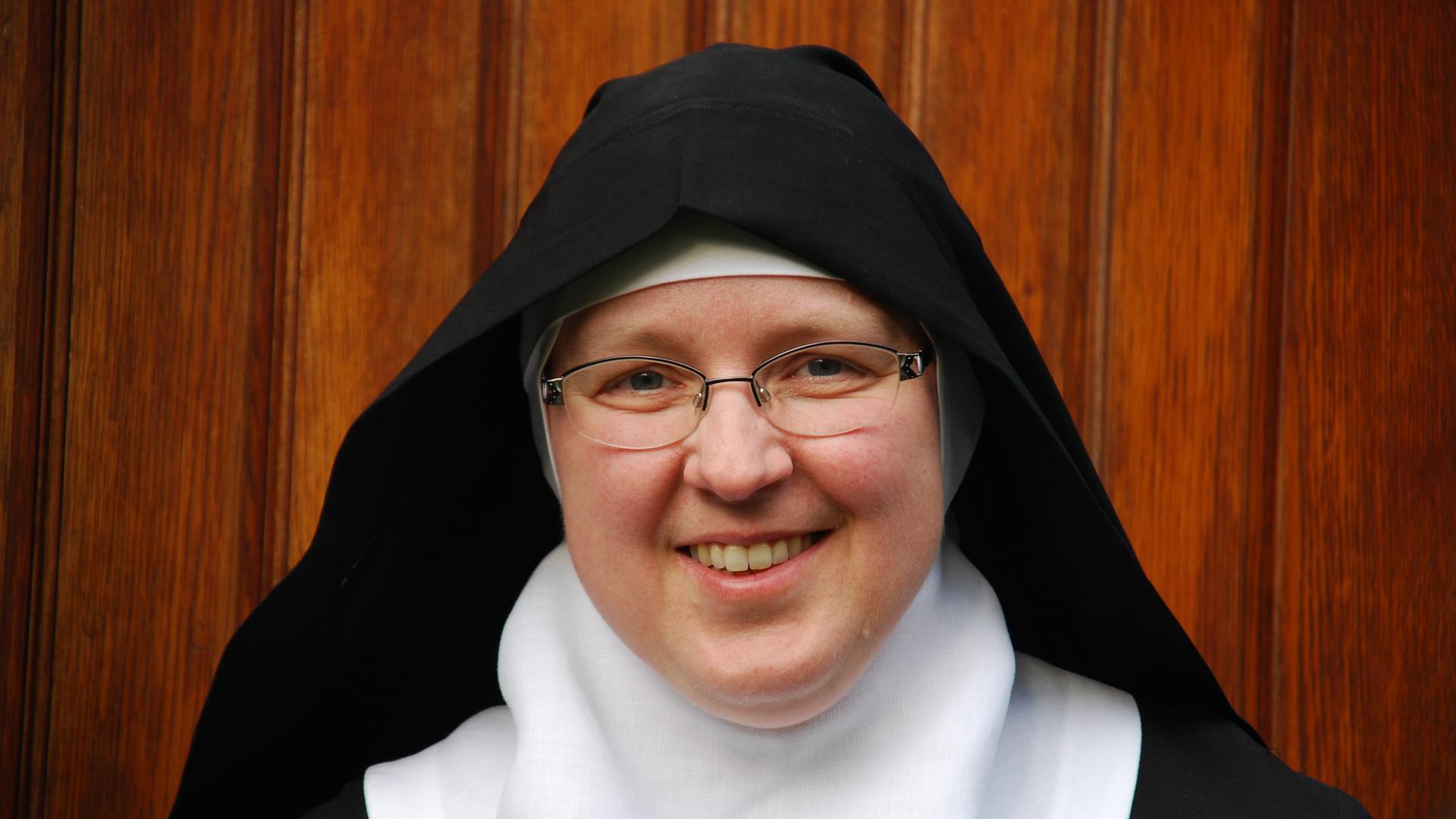 Eine Nonne in Habit lächelt in die Kamera. Im Hintergrund ist eine Wand aus Holzdielen zu sehen. Eine Nonne in Habit lächelt in die Kamera. Im Hintergrund ist eine Wand aus Holzdielen zu sehen.