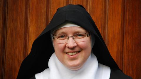 Eine Nonne in Habit lächelt in die Kamera. Im Hintergrund ist eine Wand aus Holzdielen zu sehen.